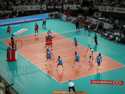 An image from an international match between Italy and Russia in 2005. A Russian player on the left has just served, with three men of his team next to the net moving to their assigned block positions from the starting ones. Two others, in the back-row positions, are preparing for defense. Italy, on the right, has three men in a line, each preparing to pass if the ball reaches him. The setter is waiting for their pass while the middle hitter with no. 10 will jump for a quick hit if the pass is good enough. Alessandro Fei (no. 14) has no passing duties and is preparing for a back-row hit on the right side of the field. Note the two liberos with different color dress. Middle hitters/blockers are commonly substituted by liberos in their back-row positions.