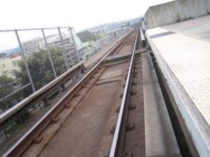 A photo of the third rails used on the BART system. Notice how the rail changes location relative to the train upon entering the station and the crossover walkway crossing the trackway.  Notice the walkway on the left side of the trackway in the distance, which is the emergency walkway for the aerial trackway leading into the Daly City station – again, the third rail positioned opposite of this walkway.