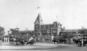 A view of the Main Street railroad depot in 1955. The building is in the Queen Anne style with mansard roofs, widow's walks, dormers, and a clock tower. People walk by a cannon, streetlamps, and a horse-drawn surrey.