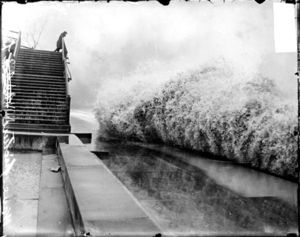A wave breaking on the shore of Lake Michigan while a man watches from a bridge.