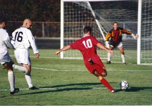  A player (wearing the red kit) has penetrated the defence (in the white kits) and is taking a shot at the goal.  The goalkeeper will attempt to stop the ball from entering the goal.