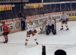 Two defencemen and a goaltender guard their goal. The referee's raised arm indicates that he intends to call a penalty.